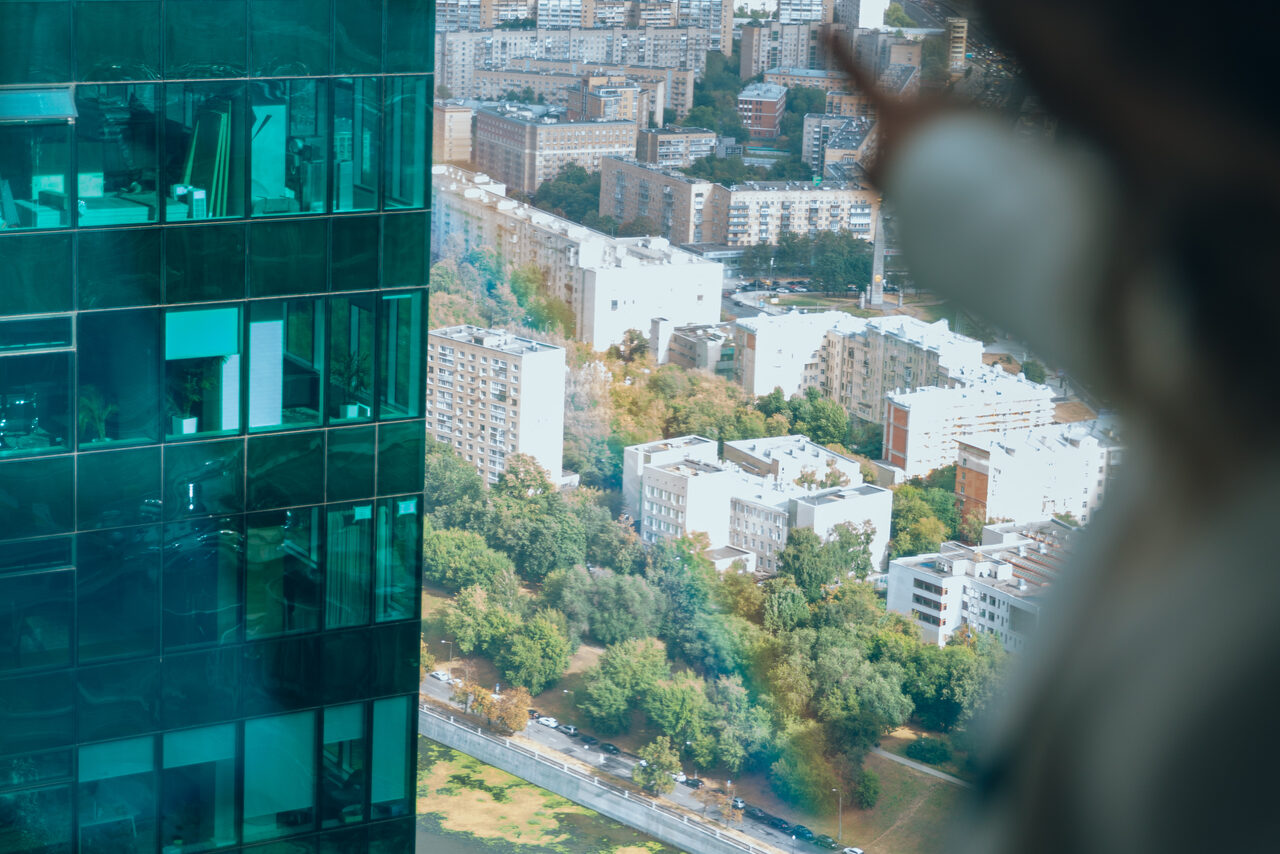 Арендовать Лекторий на 53 этаже Москва-Сити - 100 м², до 35 человек. Вид из окна высотного здания с отражением офиса на стекле. Внутри видны подсвеченные окна с офисной мебелью. За окном городской пейзаж с многоэтажными домами и деревьями. На переднем плане размытый силуэт человека.
