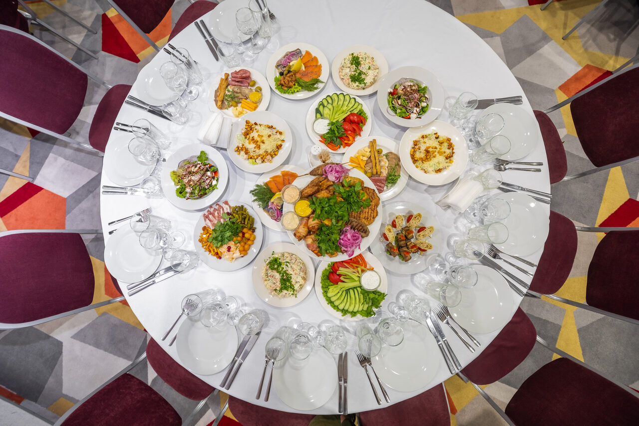 Арендовать Gorskiy City Hotel. Круглый стол, сервированный для банкета; вокруг белой скатерти расположены блюда с салатами, мясом и овощами. Для каждого гостя подготовлены тарелки, вилки и бокалы. Вокруг стола расставлены стулья с бордовыми сиденьями. На полу ковролин с геометрическим узором.