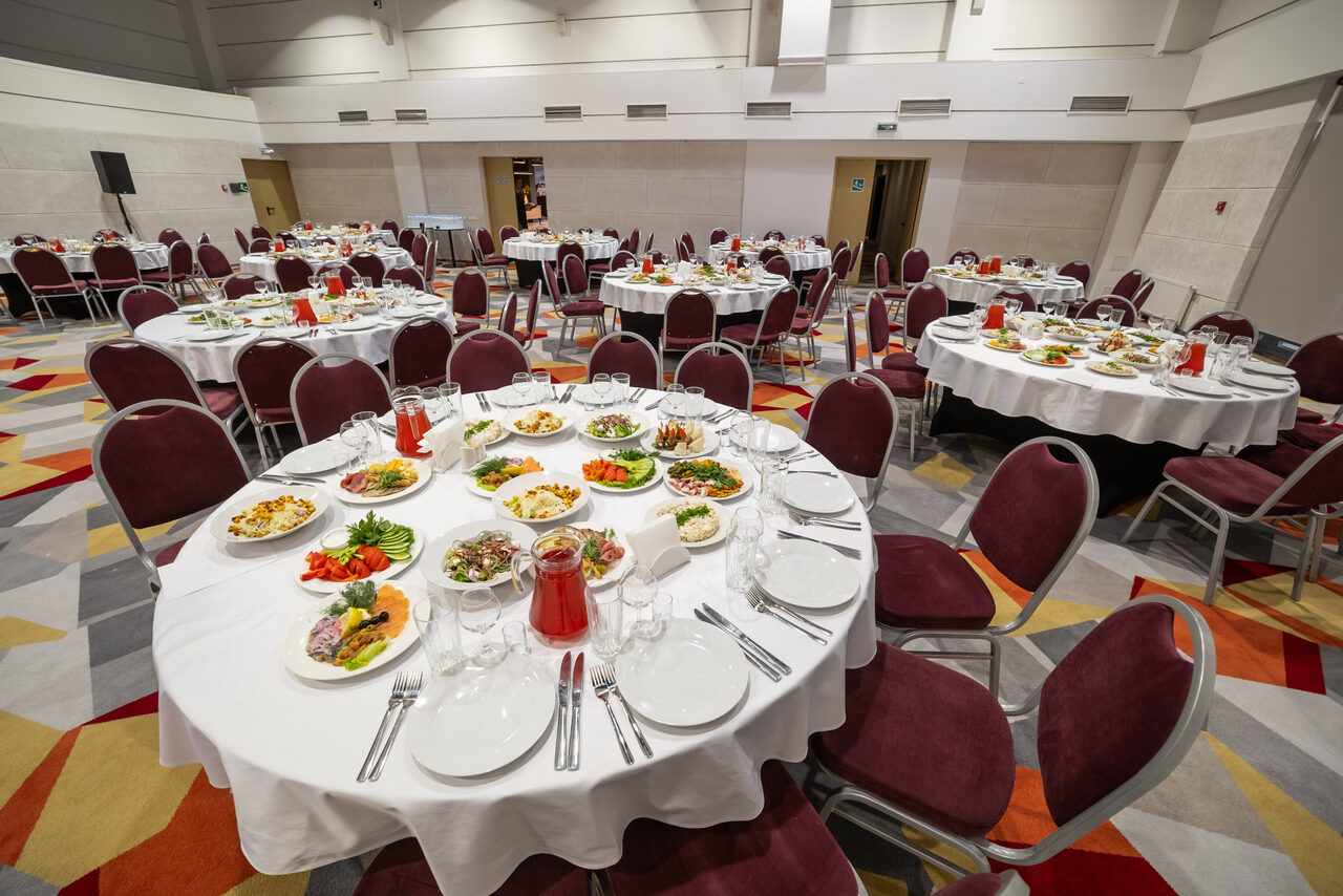 Арендовать Gorskiy City Hotel. Большой банкетный зал с круглой расстановкой столов, накрытых белыми скатертями. На столах находятся блюда с закусками, столовые приборы и стаканы. Стулья с бордовыми сиденьями, ковровое покрытие с геометрическим узором.