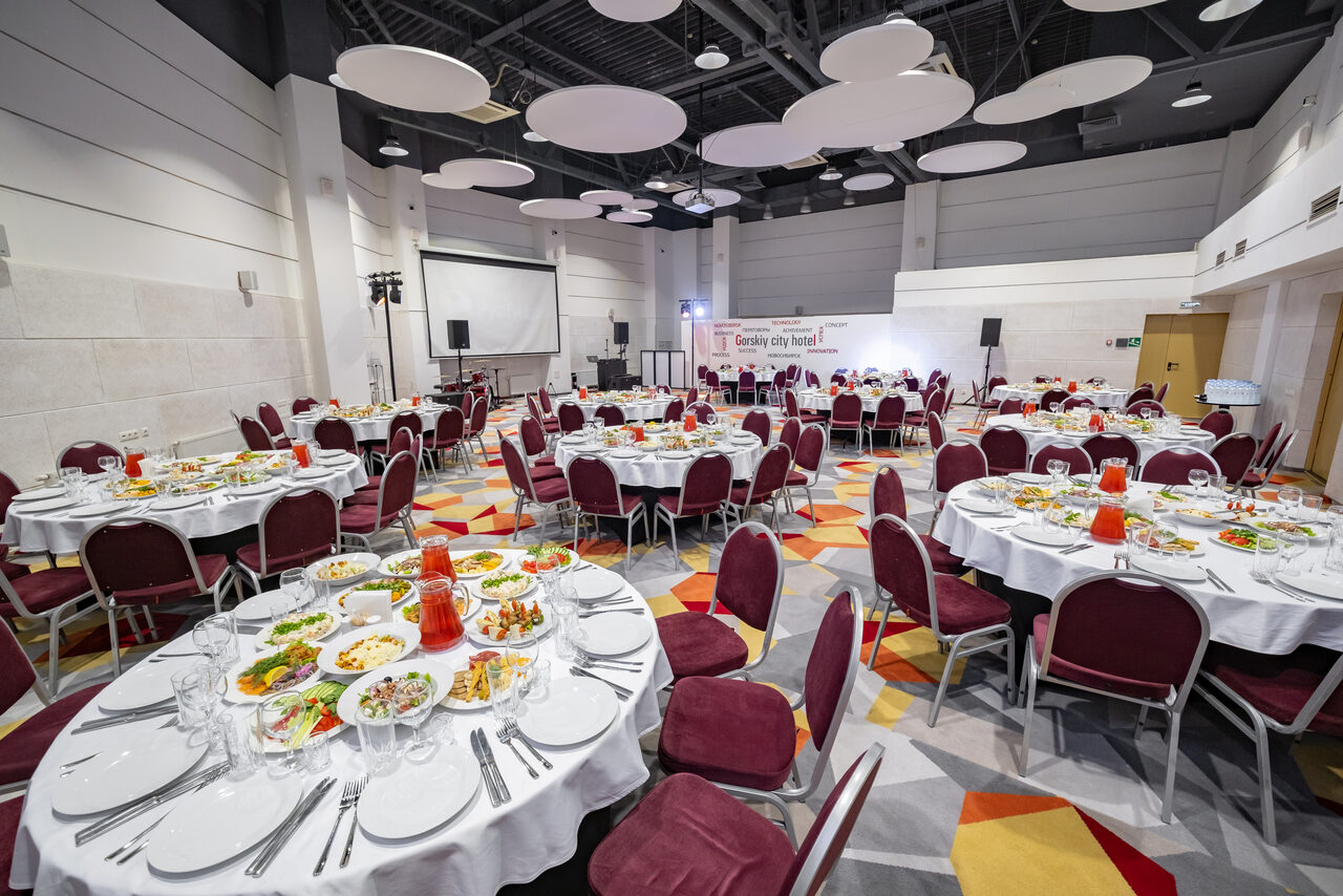 Арендовать Gorskiy City Hotel. Просторный банкетный зал с круглыми столами, покрытыми белыми скатертями, и красными стульями. На столах сервировка блюд и напитков. Потолок с круглыми подвесными элементами. Пол с геометрическим ковровым узором. Боковые стены белые.