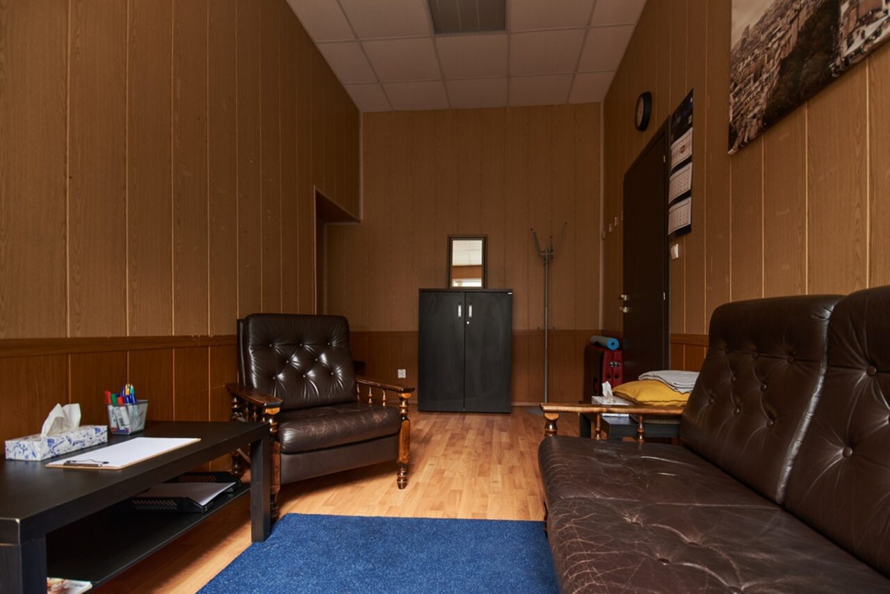 Арендовать Кабинет для работы на день или месяц . Офисное пространство с коричневыми панелями на стенах, кожаным диваном и креслом, черным столом с канцелярией, ковром синим на деревянном полу, черным шкафом, вешалкой, потолок с плиткой, дверь с расписанием, картина на стене.
