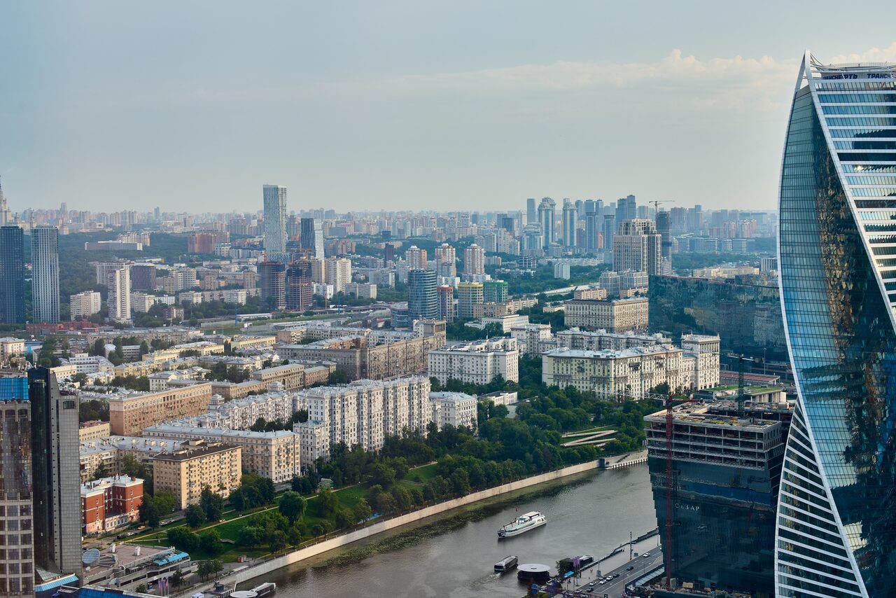Арендовать Квартира для мероприятий Свидания в Москва Сити. Вид с высоты на городской пейзаж с реками и зданиями, в том числе многоэтажки и офисные небоскрёбы. На переднем плане река с яхтой, справа современное высотное здание с изогнутым фасадом. День, облачное небо.