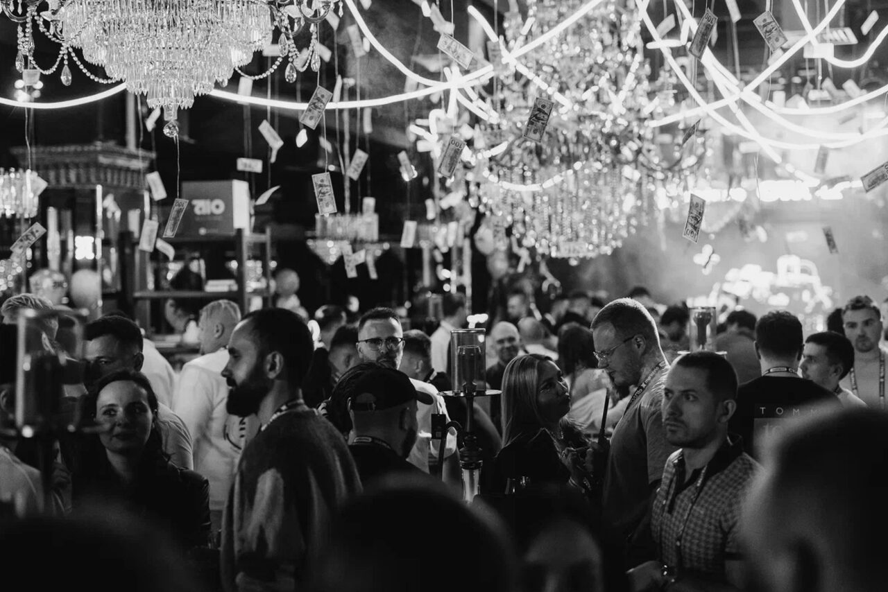 Арендовать Soul Event. Чёрно-белое изображение оживлённого мероприятия в просторном помещении. Люди общаются под люстрами и декоративными огнями. Стулья, столы отсутствуют. Надписи и знаки неразборчивы. Интерьер украшен светящимися элементами.