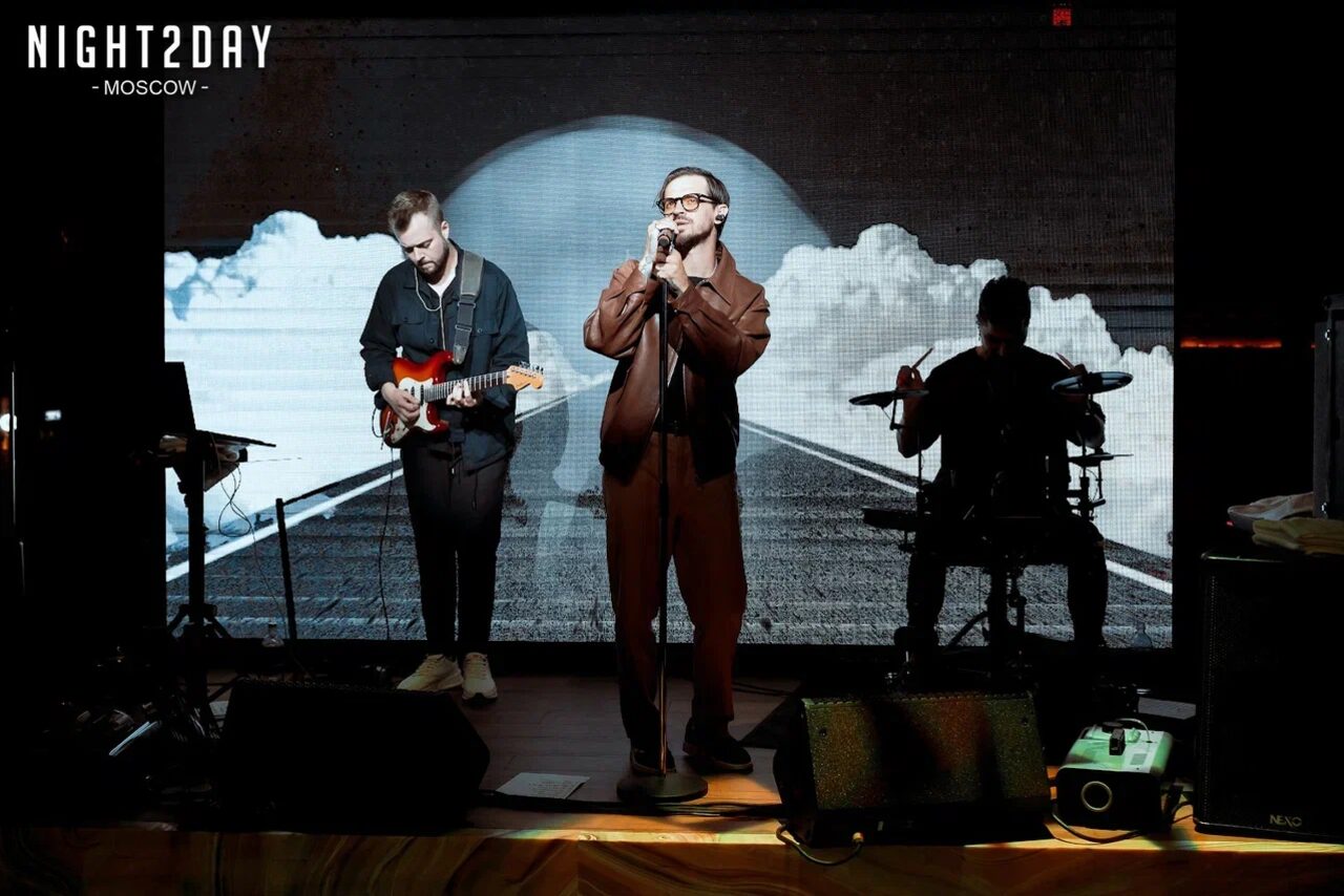 Арендовать Soul Event. Музыкальная сцена с тремя музыкантами: один с гитарой, второй с микрофоном, третий за ударными. Позади них экран с изображением дороги и облаков. Освещение направлено на музыкантов. На полу акустическое оборудование.