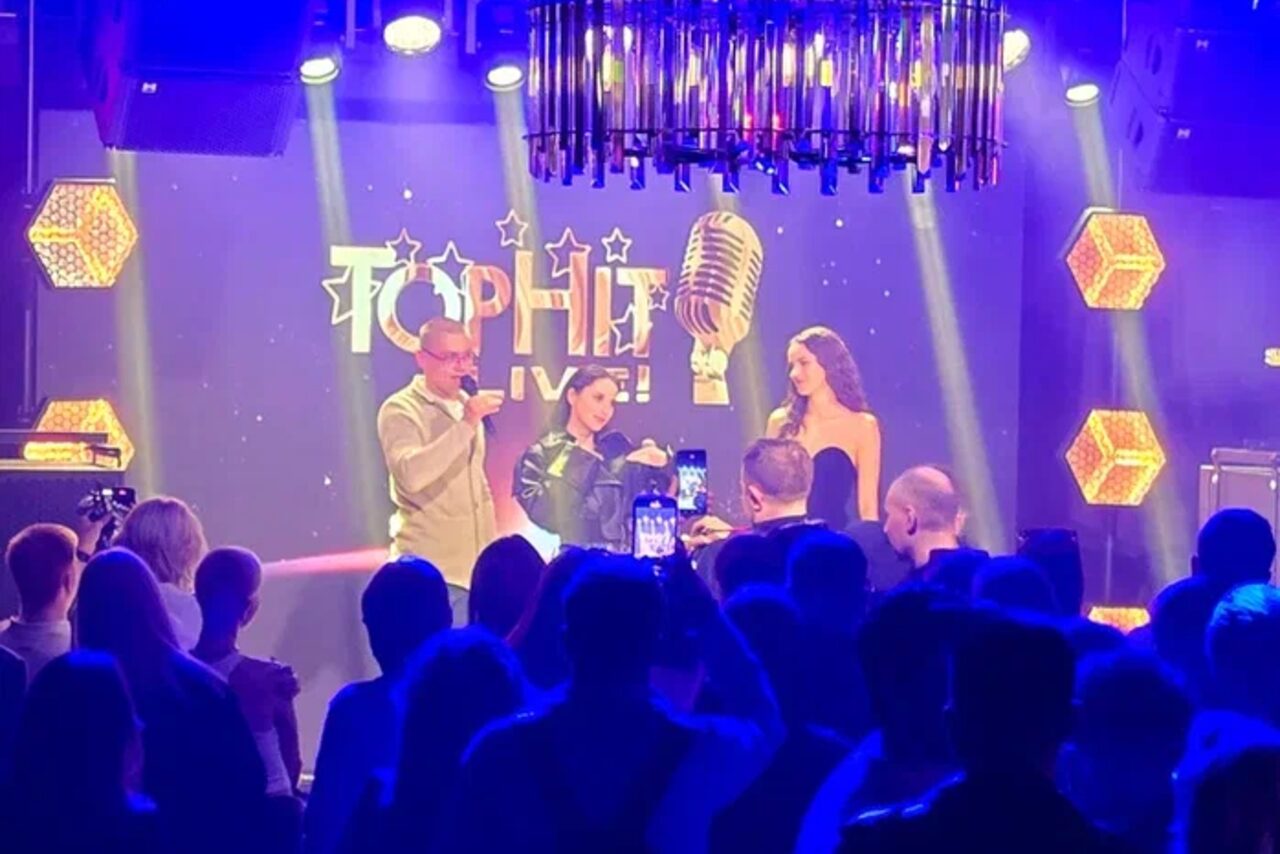 Арендовать Soul Event. Люди на сцене в помещении с приглушенным освещением, металлическая люстра. На заднем плане экран с микрофоном и текстом "TOPHIT LIVE". По бокам светящиеся шестиугольные панели. Перед сценой сидит или стоит публика.