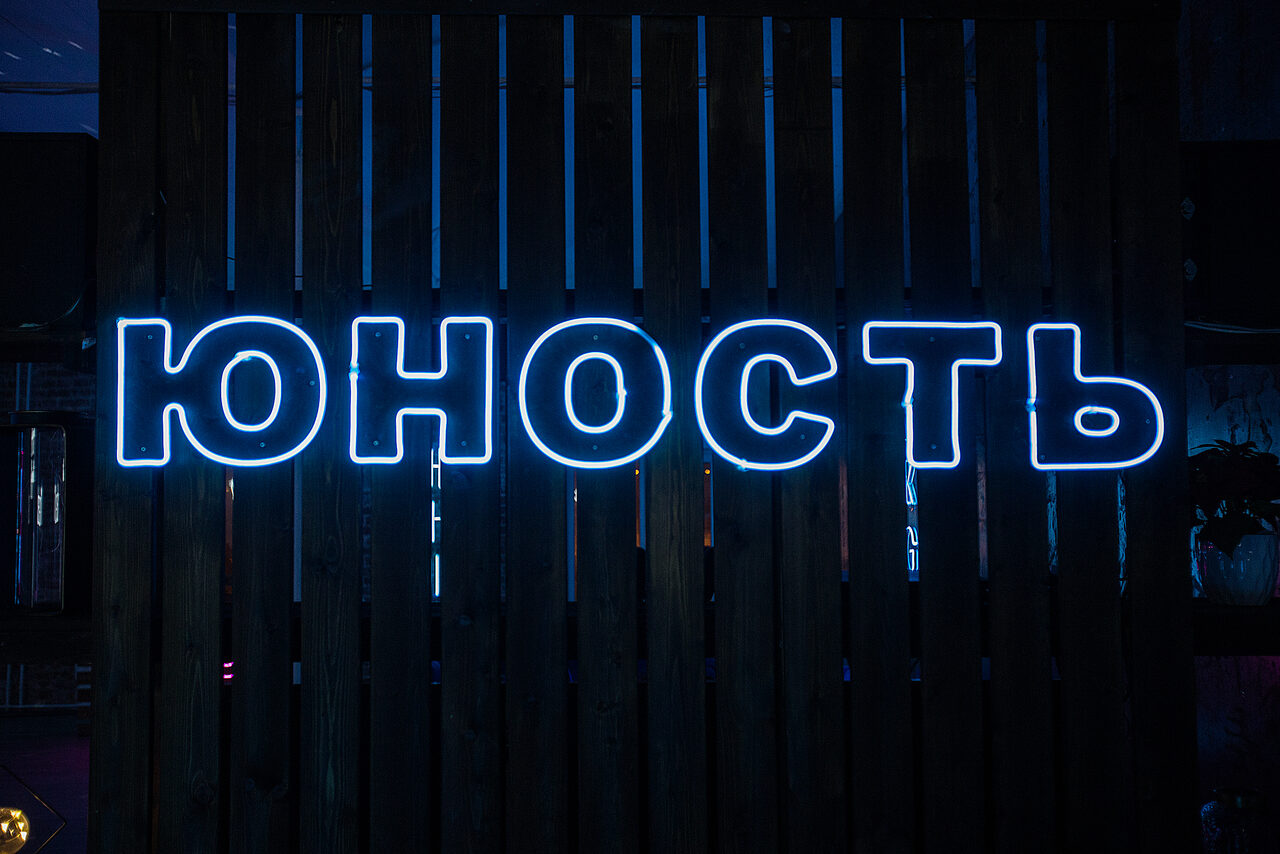 Арендовать ЮНОСТЬ - Стильный и уютный лофт для любого мероприятия. Топ-сервис!. Неоновая вывеска с надписью "ЮНОСТЬ" на деревянной стене, подсвеченная голубым светом. Помещение затемнено, и другие детали видны слабо.