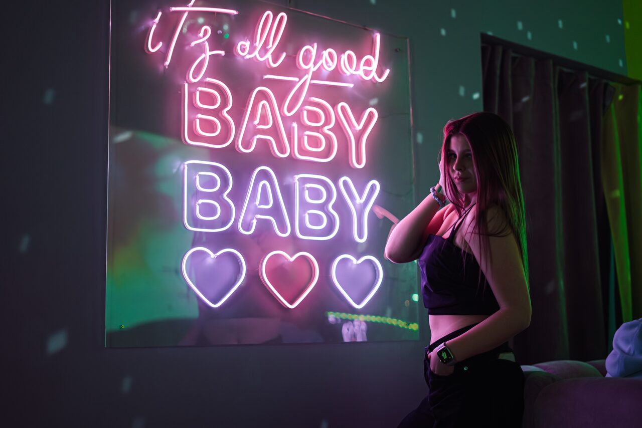 Арендовать Лофт ЗЕФИРКА. Неоновая вывеска с фразой "1/3 all good BABY BABY" и тремя сердцами на тёмной стене. Справа находится человек возле занавески и мягкой мебели. Обстановка с приглушенным освещением.