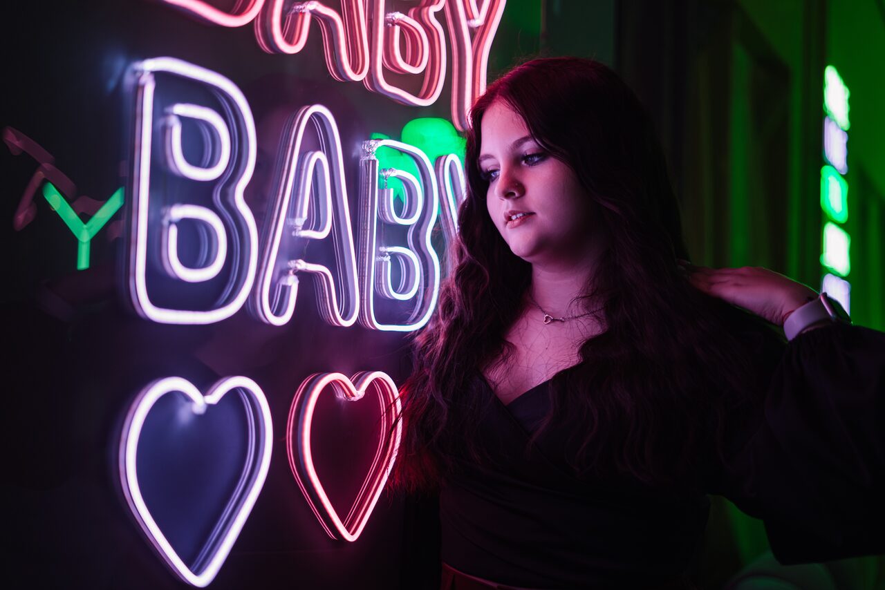 Арендовать Лофт ЗЕФИРКА. Неоновая вывеска с текстом "BABY" и сердцами, светящаяся белым и розовым цветами на фоне темного помещения. Освещение в зелено-фиолетовых тонах, девушка в черной одежде стоит возле стены.