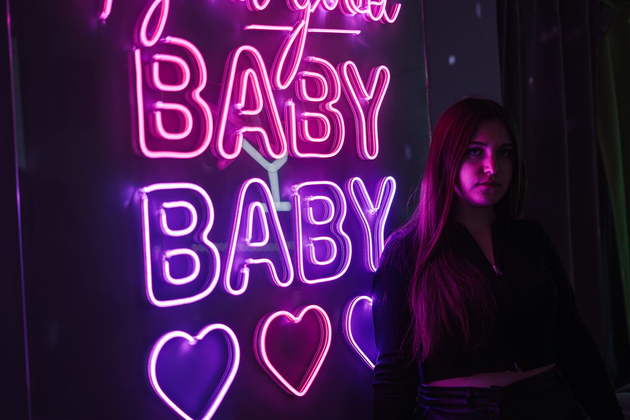 Арендовать Лофт ЗЕФИРКА. Неоновая вывеска с повторяющимся словом "BABY" и сердцами, светящаяся розовым и фиолетовым цветами. Женщина стоит рядом с вывеской. Темное окружение, акцент на освещении.