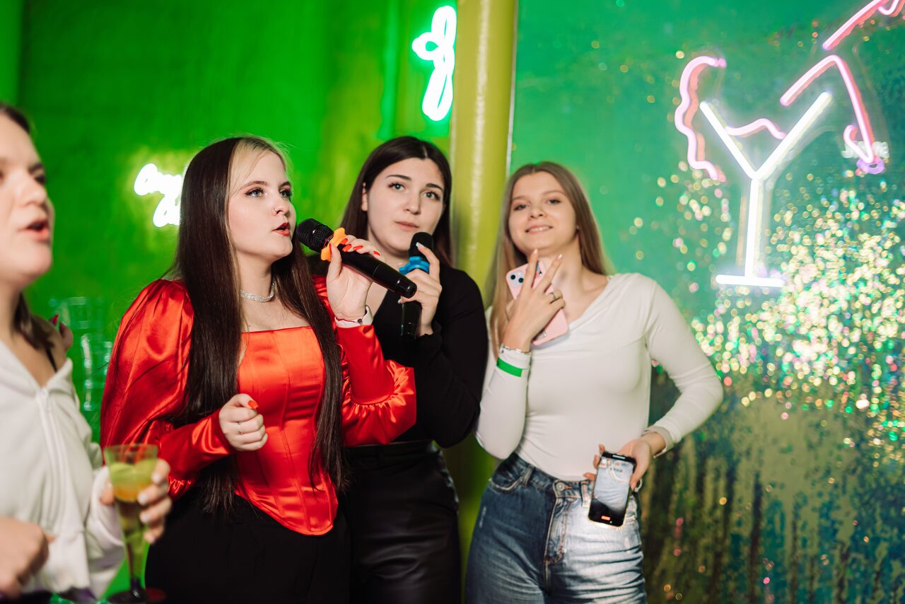 Арендовать Лофт ЗЕФИРКА. Женщины поют в микрофон в помещении с зелеными и блестящими стенами. На заднем плане видны неоновые подсветки в виде бокала. Женщины одеты в яркую одежду, одна держит смартфон. Освещение мягкое с разноцветными акцентами.
