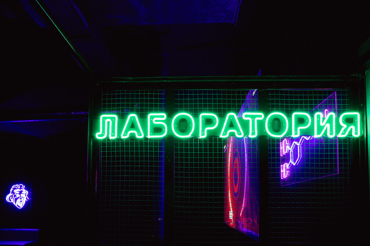 Арендовать ЛАБОРАТОРИЯ - необычный лофт для тематических мероприятий. Лучший сервис, классный звук, приставка и лед в подарок!. Неоновая надпись "ЛАБОРАТОРИЯ" на черной проволочной сетке, освещенная зеленым светом. На заднем плане видны элементы фиолетового и красного неона. Темное помещение, оформленное в стиле с использованием сеток и неоновых вывесок.