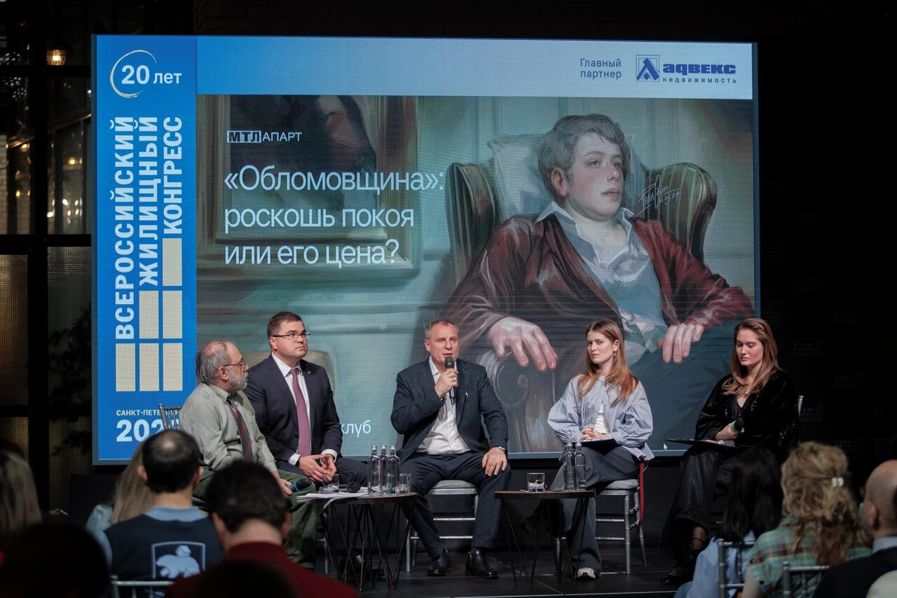 Арендовать Банкетное пространство ресторана площадью 600 м 2 . Конференц-зал с пятью людьми на сцене, сидящими на стульях перед аудиторией. Большой экран с изображением и текстом позади них. Серые стены, черный потолок, экран с синими и белыми элементами. Люди одеты в деловой и повседневный стиль.