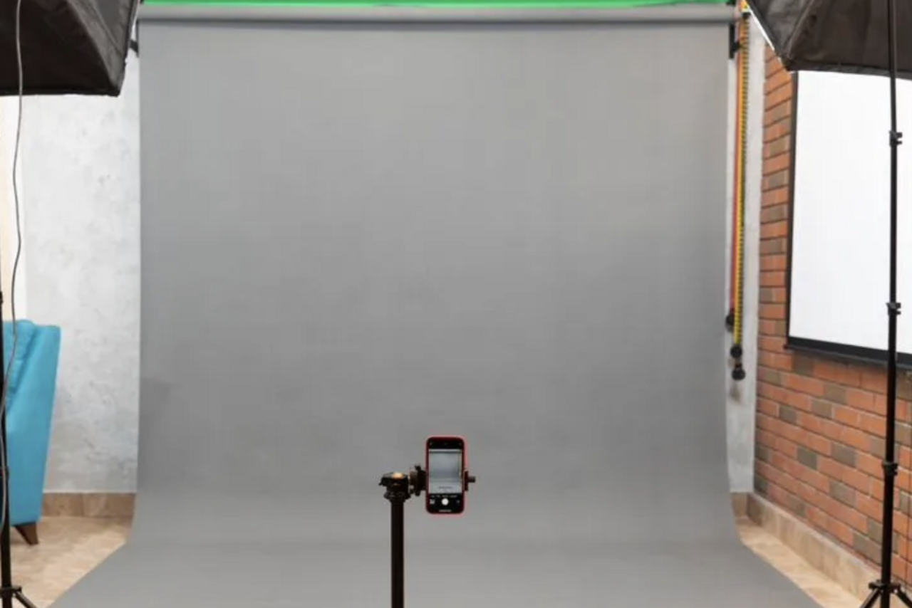 Арендовать Светлая фотостудия. Фотостудия с серым фоном, двумя студийными светильниками слева и справа, красно-кирпичной стеной справа и белой стеной слева. В центре расположен штатив с прикрепленным смартфоном. На полу светлый кафель.