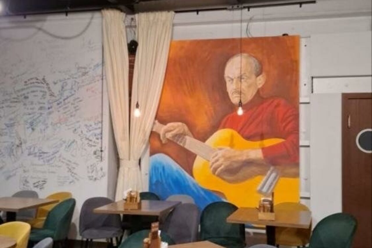 Арендовать Гнездо Глухаря. Кафе или пространство для встреч с деревянными столами и разноцветными стульями. На стене большое изображение человека с гитарой. Светлые занавески отделяют зону. Подвесные лампы создают мягкое освещение. На стене виден текст.