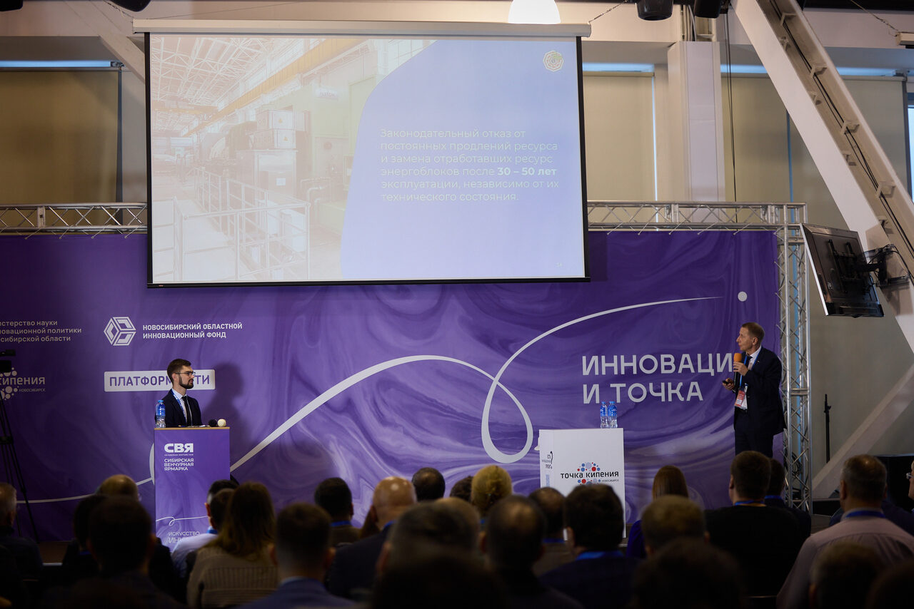 Арендовать Точка кипения - Новосибирск. Аудитория собралась в конференц-зале. Два человека на сцене перед экраном. Фиолетовый фон с текстами. Люди сидят и смотрят презентацию. Проекторы, микрофоны и располагающийся спереди экран с изображением зала.