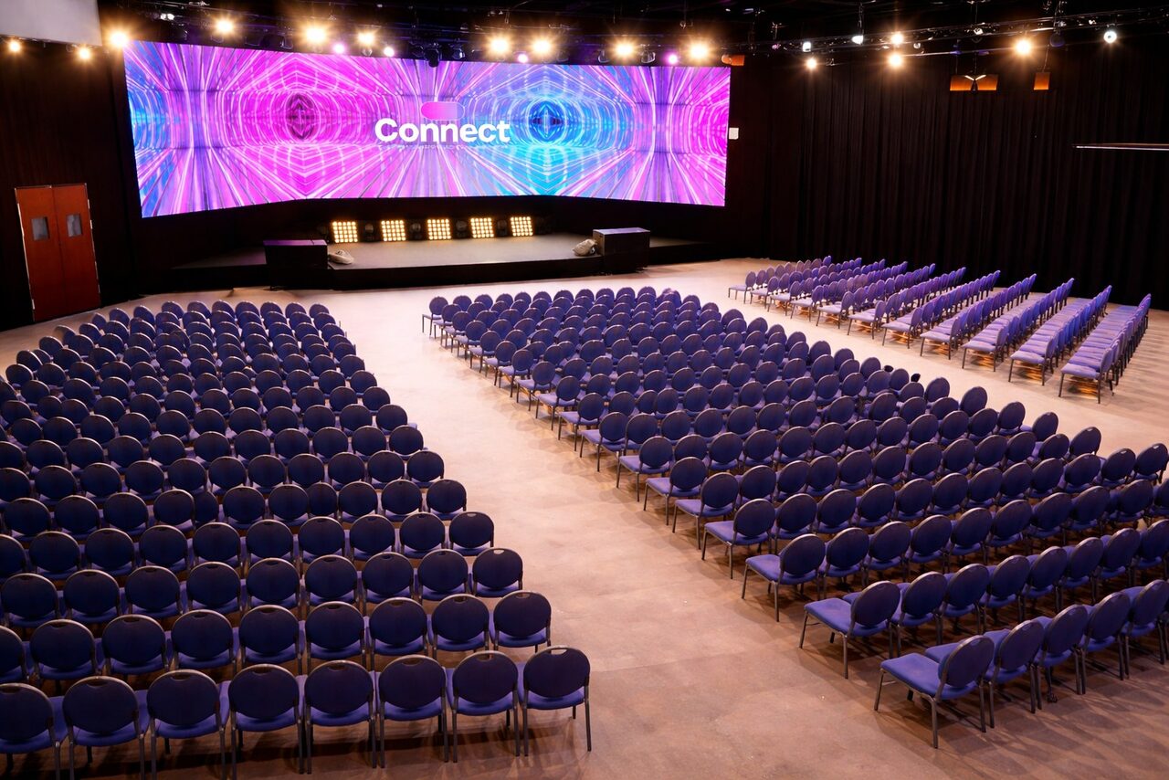 Арендовать Connect — конгресс-центр на 1000 гостей. Большое помещение с конференц-залом, вмещающим ряды синих стульев. На сцене расположены кресла, за ней — широкий экран с текстом "Connect" и абстрактным рисунком фиолетового и синего цветов. Светильники на потолке. Пол светлый.