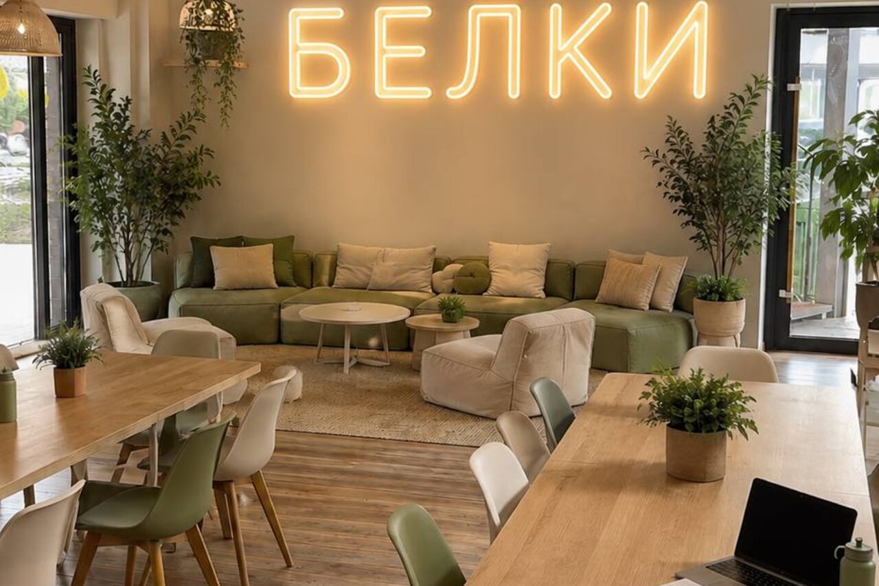 Арендовать БЕЛКИ. Просторное помещение с деревянными столами и стульями, зеленый диван с подушками, кресла, растения в горшках. На стене неоновая надпись. Большие окна, светлые стены, лампы и естественное освещение.