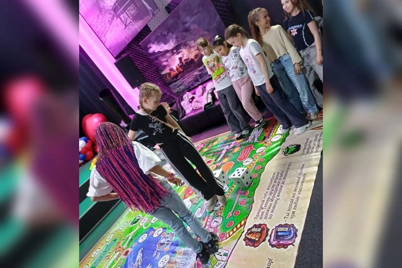 Арендовать Оборудованная площадка в лофт стиле. Детская игровая комната с ярким напольным покрытием, изображающим игровое поле с текстом и изображениями, дети стоят по кругу. На заднем плане мягкая мебель и стены с фиолетовым освещением и картинами.