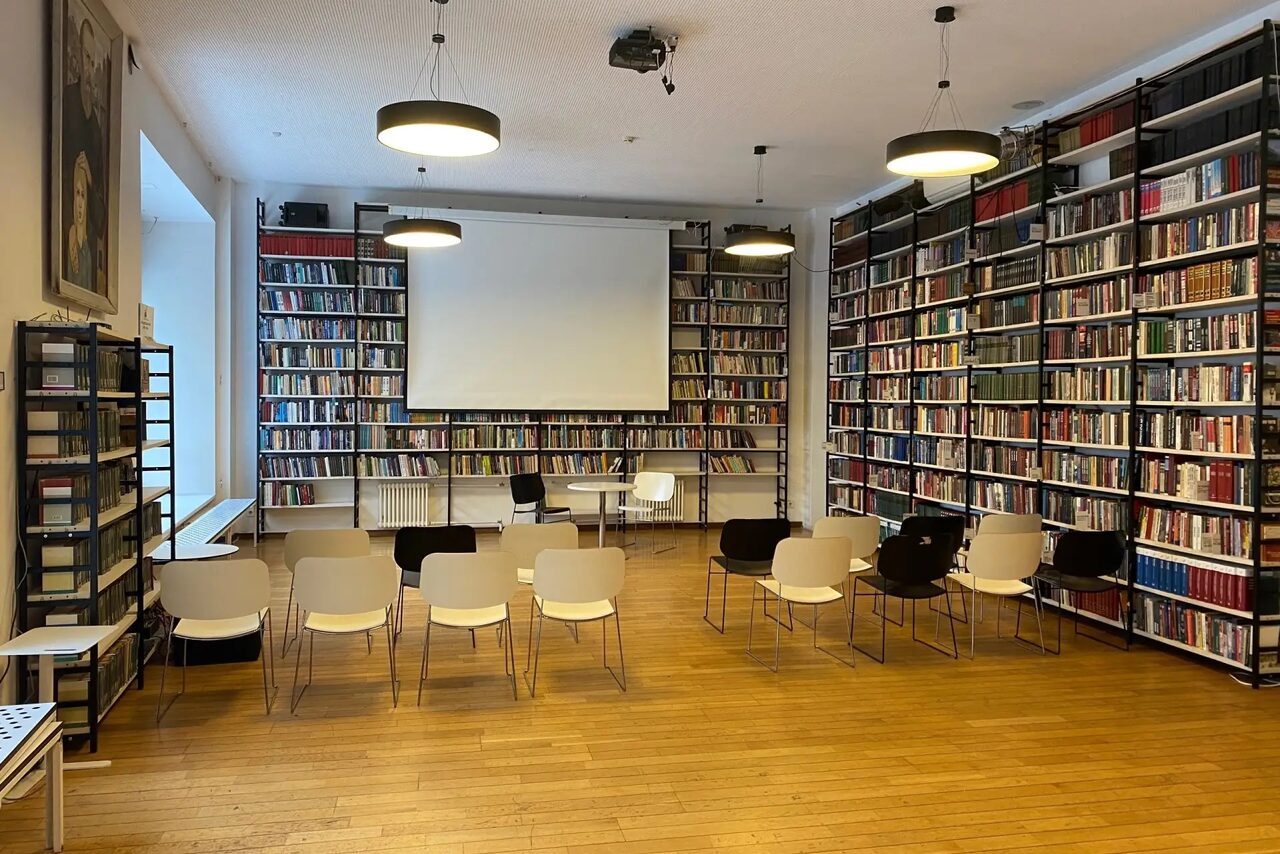 Арендовать Зал для вашего мероприятия. Помещение библиотеки с книжными стеллажами вдоль стен, проекторным экраном впереди. Расставлены стулья в несколько рядов. Потолочные светильники, светлый деревянный пол, белые стены. Видно большое окно слева.