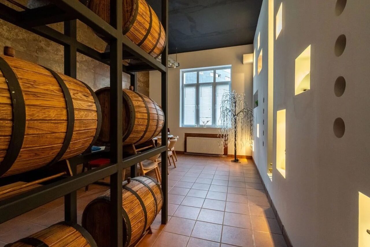 Арендовать La Cueva room. Пространство с деревянными бочками, стеллажами, столом и стульями. Терракотовая плитка на полу, белые стены с квадратными нишами, большое окно. Освещение встроенное и естественное. Потолок темного цвета.