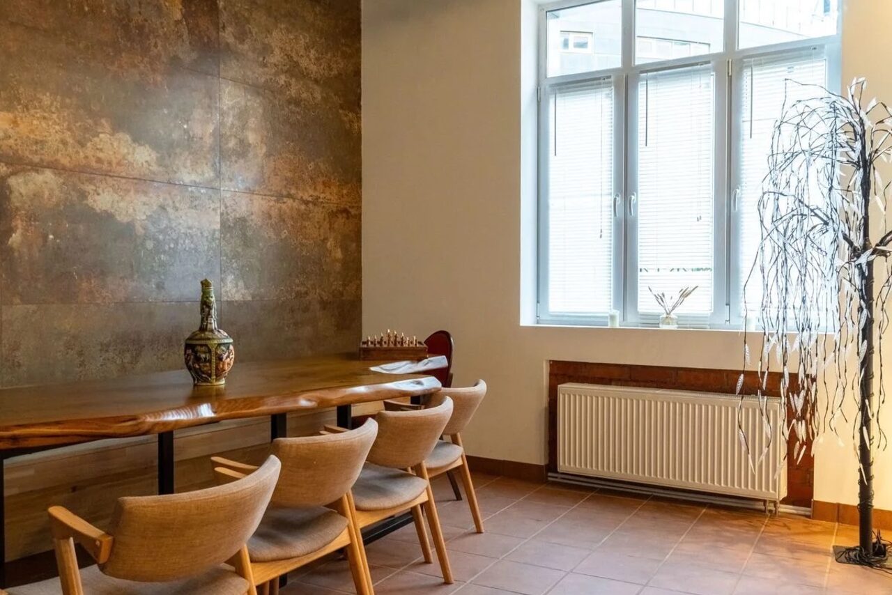 Арендовать La Cueva room. Просторная комната с деревянным столом и четырьмя стульями, стены с текстурой под бетон, большие окна с жалюзи, радиатор, декоративное дерево, пол из плитки коричневого цвета, ваза на столе.