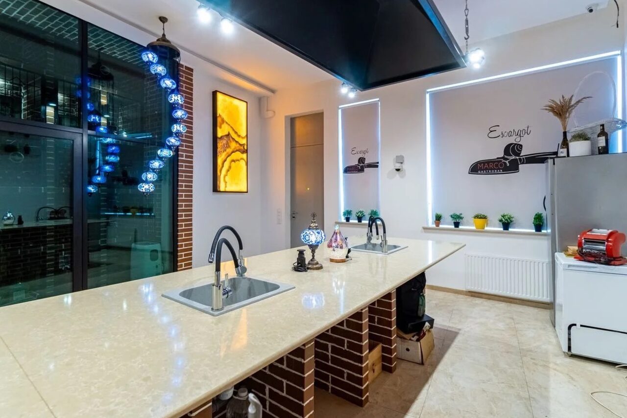 Арендовать La Cueva room. Кухонное пространство с длинным островом и двумя встроенными раковинами. Окружено кирпичными панелями и подвесными светильниками с синими элементами. На стене декор в виде панно и светодиодной ленты. Есть холодильник, декор и растения.