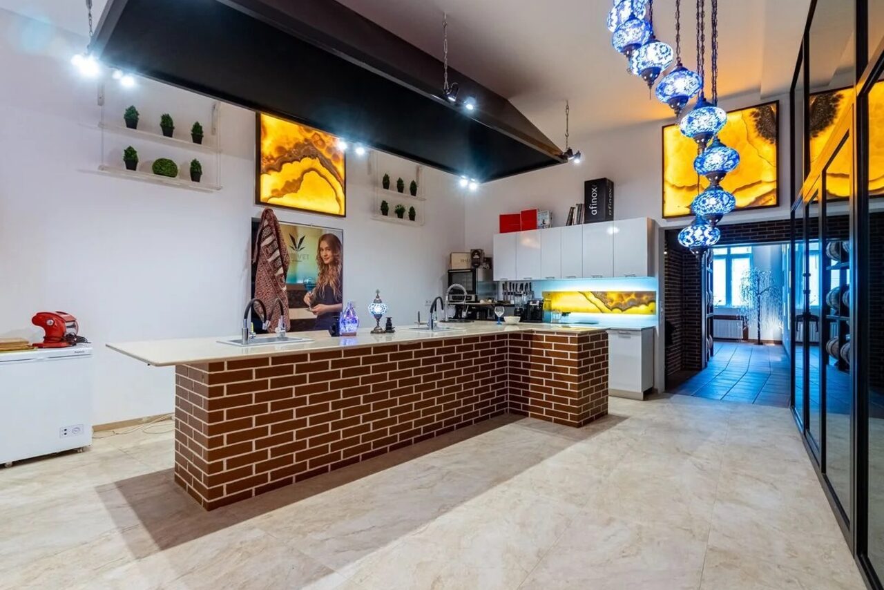 Арендовать La Cueva room. Кухонное пространство с островом, облицованным кирпичом. Белые шкафы, встроенные светильники, декоративные панели с подсветкой. Подвесные лампы синих оттенков. Плиточный пол, светлые стены. Видны кухонные приборы и декор.