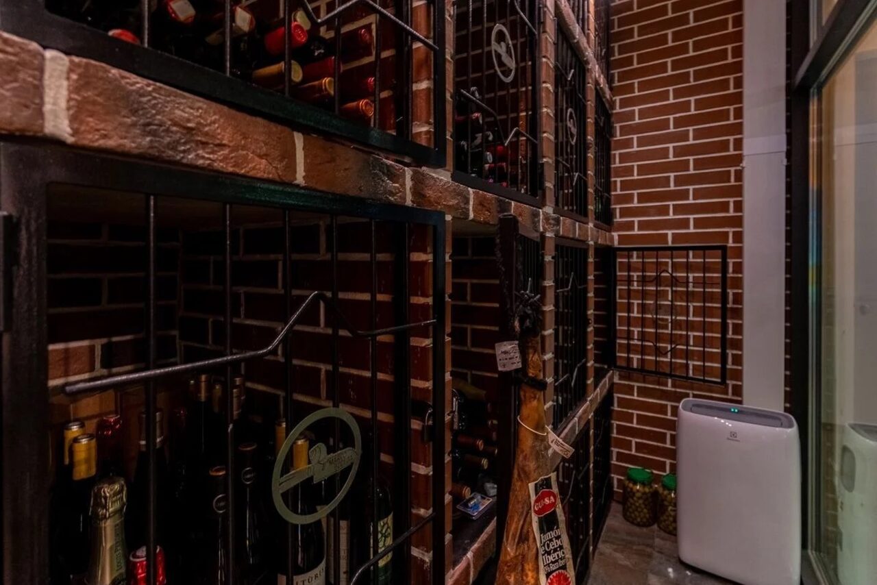 Арендовать La Cueva room. Помещение с кирпичными стенами и винными стеллажами из металла. На полках видны бутылки вина за решетками. Справа стоит портативный кондиционер. Освещение приглушенное. Видна надпись на нечеткой черной табличке.