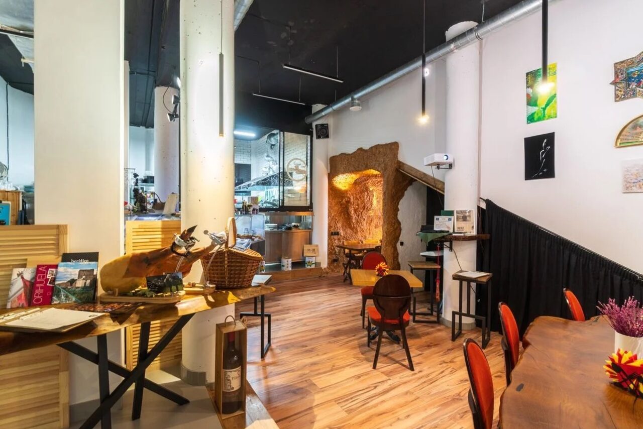 Арендовать La Cueva room. Кафе с деревянным полом и высокими потолками. Зона с мягкими стульями и деревянным столом, декорирована камином. Освещение люстрами и светильниками. Светлые стены, картины и фотографии на стенах, журналы на столе.