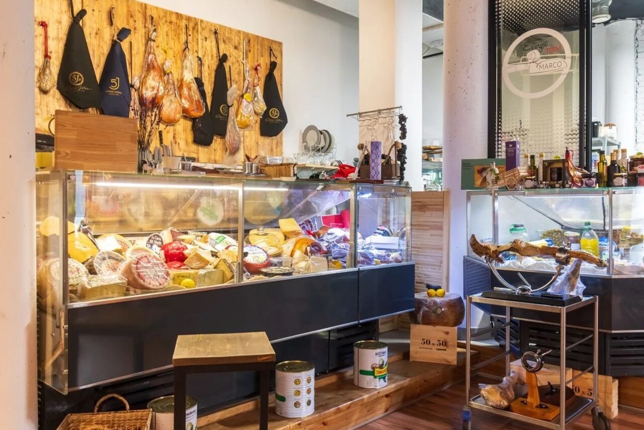 Арендовать La Cueva room. Магазин с продуктами: витрины с различными сырами и мясными изделиями, деревянная отделка, белые колонны, пол из светлого дерева, на стене деревянная панель с продуктами и черными передниками. Освещение верхнее, яркое.