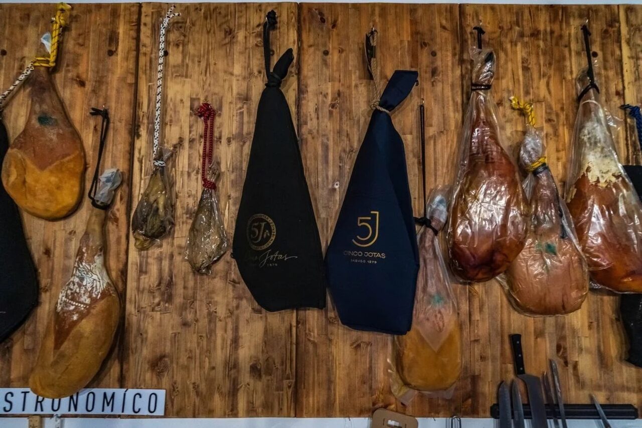 Арендовать La Cueva room. Деревянная стена с подвешенными окороками и колбасами в защитных чехлах и без, левая часть содержит видимую часть фразы "STRONOMICO".