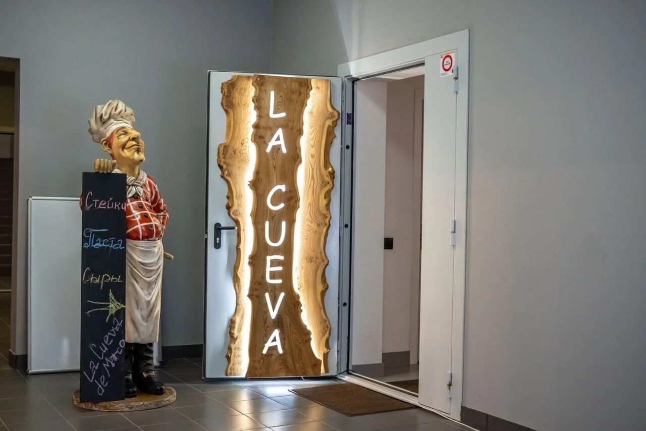 Арендовать La Cueva room. Дверной проем с открытой дверью, на двери деревянная табличка с надписью "LA CUEVA". Справа статуя шеф-повара с деревянной доской, на доске написано мелом: "Стейки", "Паста", "Сыр". Интерьер в серых тонах, пол покрыт плиткой.