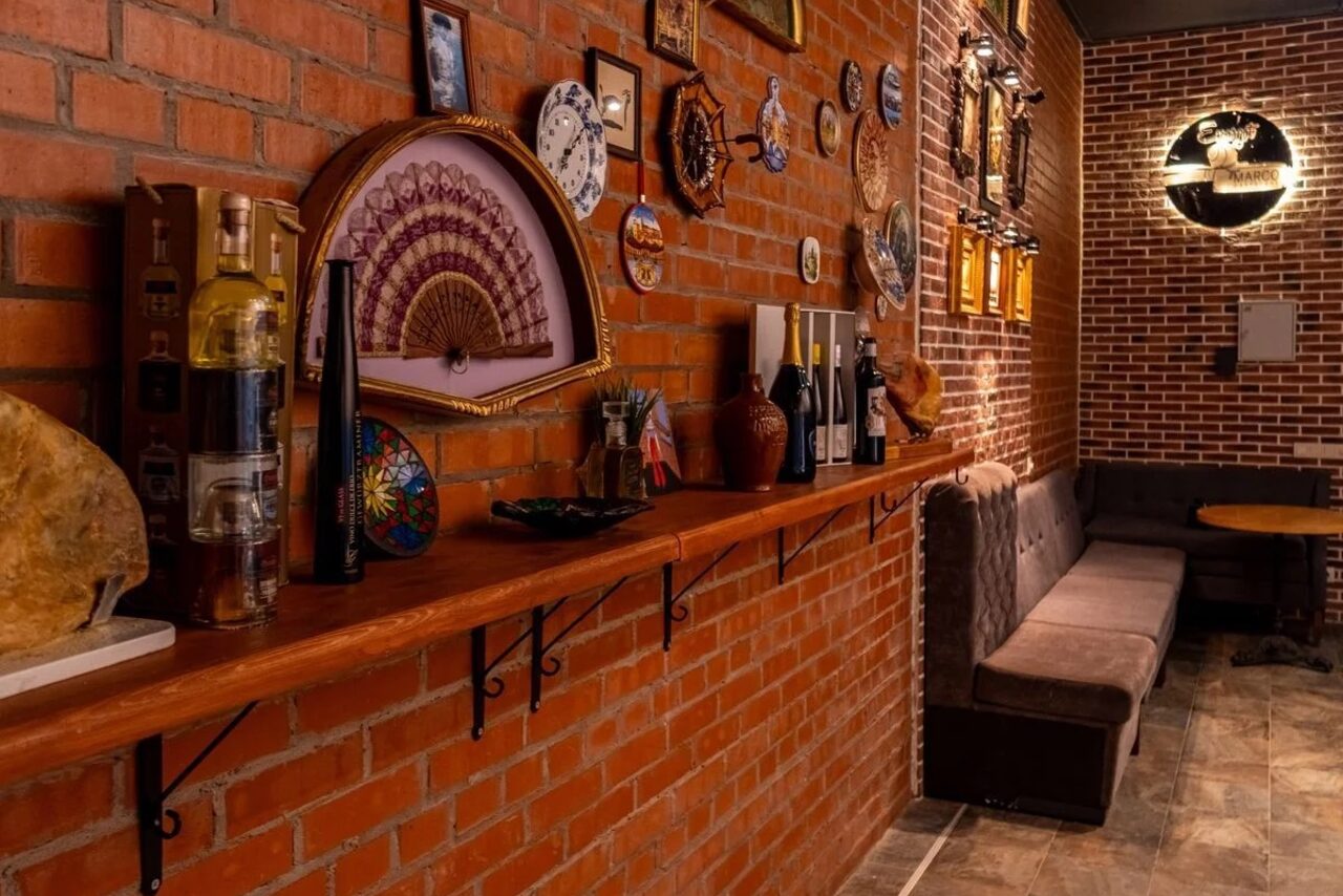 Арендовать La Cueva room. Пространство с кирпичными стенами, стеллаж с бутылками и декоративными элементами, мягкий диван вдоль стены, круглый стол, настенное освещение. Декор включает различные настенные тарелки и картины. Пол выложен плиткой.