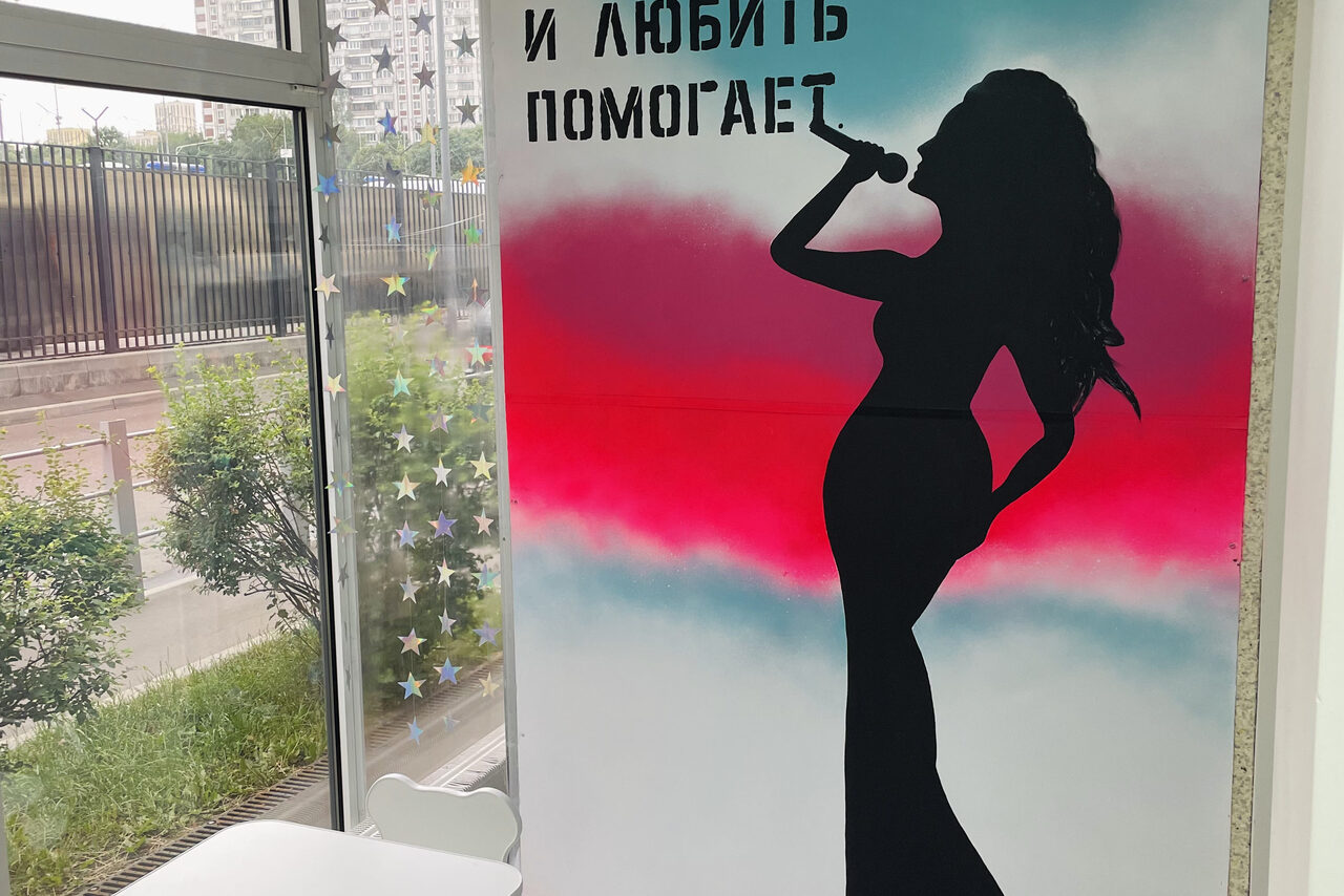 Арендовать Гармония. Пространство с панорамными окнами. На стене изображение силуэта певицы с микрофоном с разноцветным фоном. Рядом белый стол и стул. Окно украшено звездами. Снаружи зелень и забор. Надпись на стене: "И жить помогает".