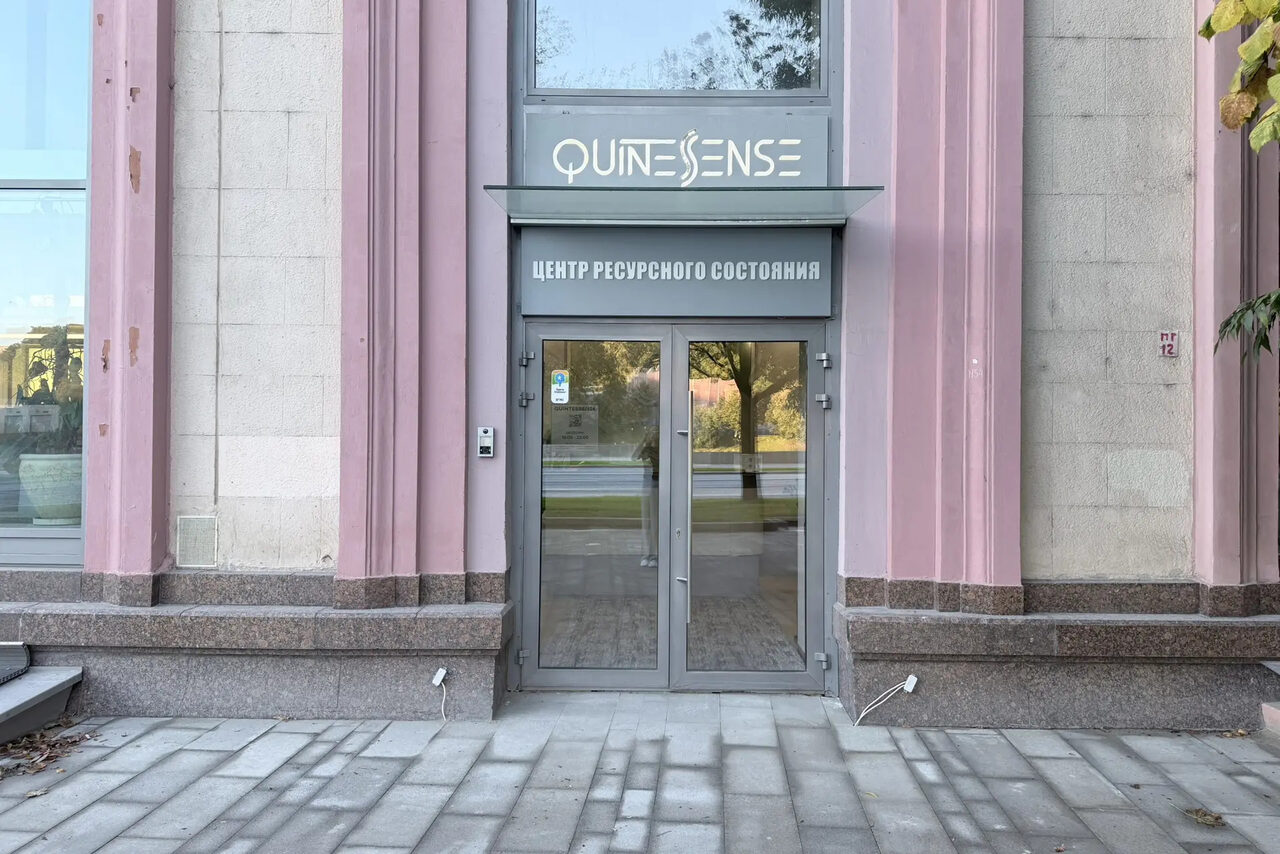 Арендовать QuintesSense. Вход в здание с двойными стеклянными дверями, серыми рамами. Над дверями вывеска с двумя текстовыми строками, верхняя содержит неразборчивый текст, нижняя — "Центр Ресурсного Состояния". Стены светло-серого цвета с розовыми колоннами.
