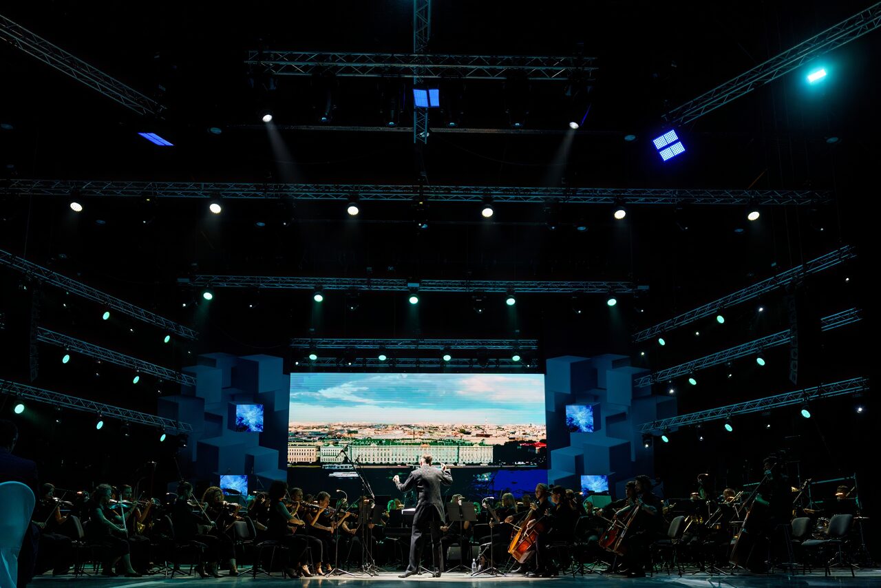 Арендовать Современный комплекс. Большая сцена с оркестром, оркестранты рассажены полукругом, дирижёр в центре. Сзади крупный экран с городским пейзажем. Освещение яркое направленное, синие и белые прожекторы. Подвесные конструкции и колонки по сторонам.