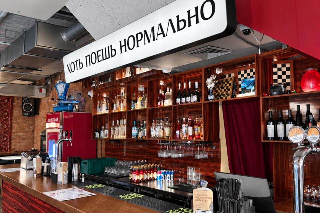 Арендовать Караоке-рюмочная Шотский. Барное пространство с длинной деревянной стойкой, оборудованной пивными кранами, стеклянная полка с алкоголем на заднем плане, яркое освещение, кирпичная стена, металлические вентиляционные трубы на потолке, световая вывеска с текстом на русском языке.
