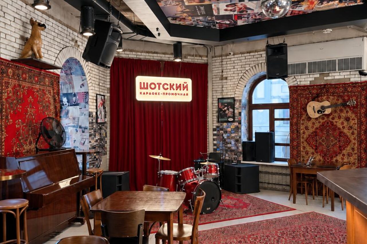Арендовать Караоке-рюмочная Шотский. Караоке-пространство с кирпичными стенами, барабанной установкой, пианино. Красные ковры, занавес, деревянная мебель, настенные декорации. Освещение включает потолочные светильники. Есть вывеска с надписью на русском языке.