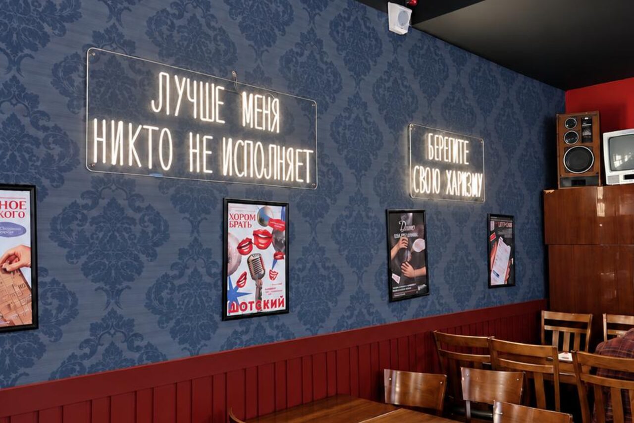 Арендовать Караоке-рюмочная Шотский. Столовая зона с деревянными стульями и столами. Синяя стена с дамасским узором, украшенная неоновыми надписями и постерами. Деревянная панель на стене, аудиоколонки сверху. Освещение дает мягкий свет.