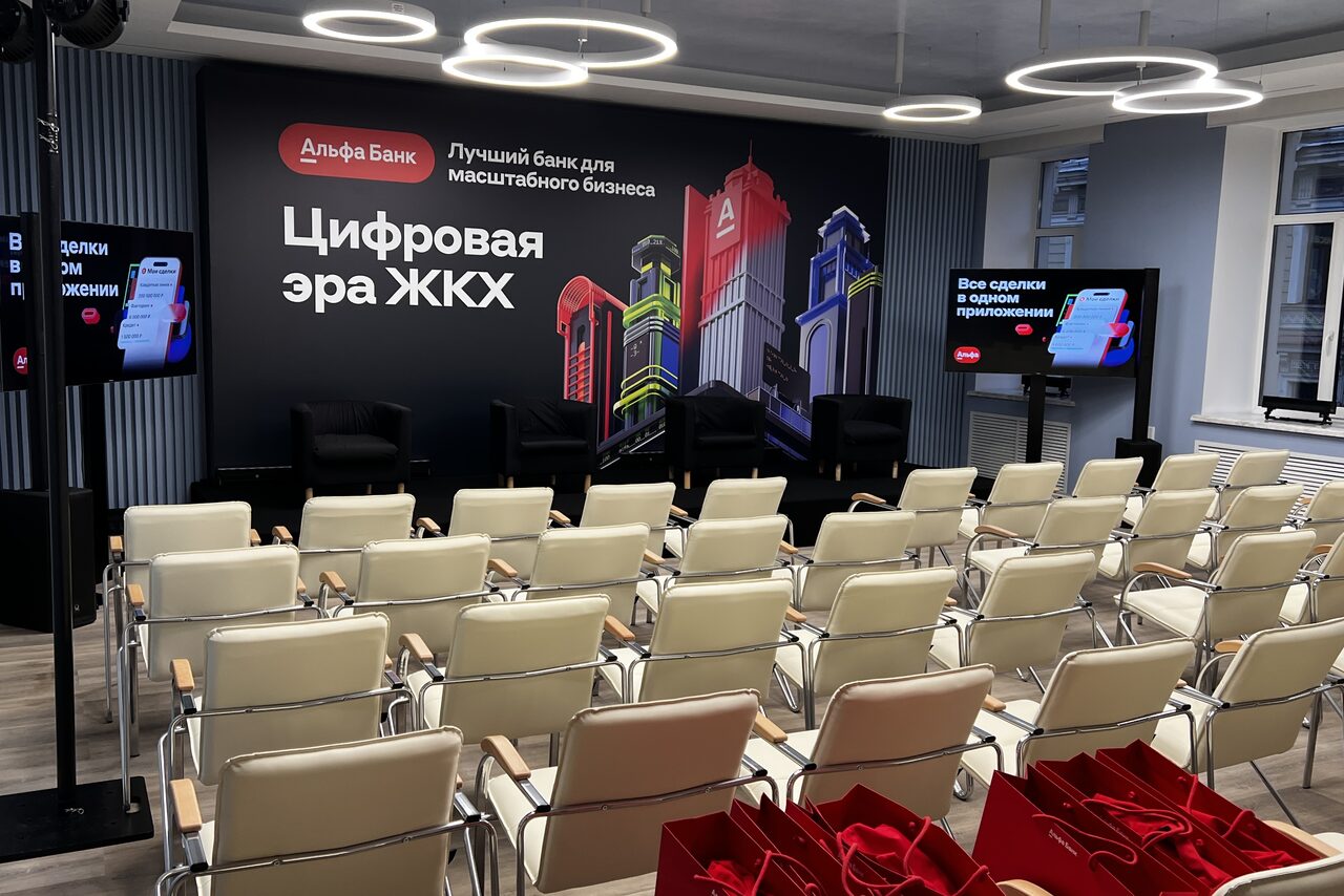 Арендовать Дом Жилищного Работника. Аудиторное пространство с рядами белых стульев, черной сценой с креслами, задником с текстом и изображениями зданий. На стенах светодиодные экраны, один с синим интерфейсом. Круглые светильники на потолке, серые стены в полоску. Красные пакеты на полу.