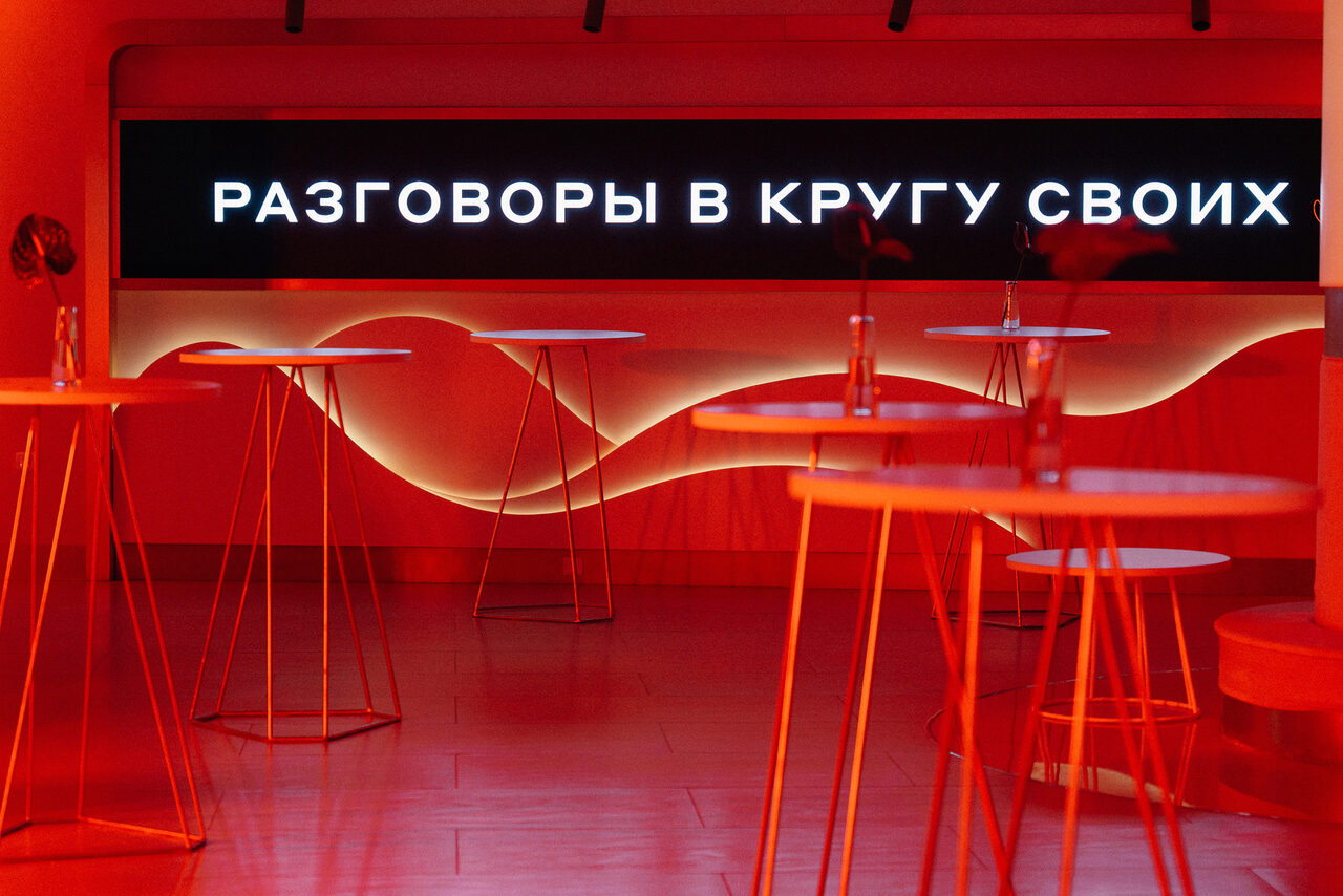 Арендовать Горький Холл. Зал с высокими металлическими столами на тонких ножках, красное освещение. На стене черная панель со светящимся текстом «РАЗГОВОРЫ В КРУГУ СВОИХ». Деревянный пол. Стены украшены волнистыми элементами.