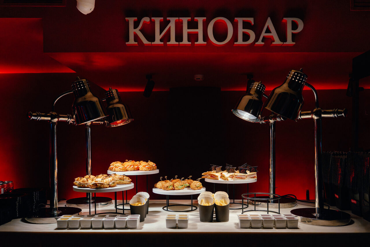 Арендовать Горький Холл. Кинобар с темно-красной подсветкой, на столе под лампами расположены подносы с выпечкой и десертами. Вывеска "КИНОБАР" на стене. Белый стол с контейнерами и черными декоративными элементами.