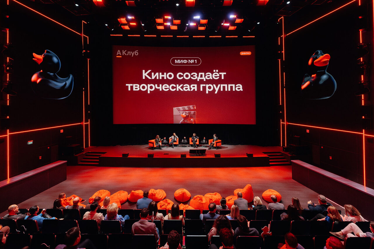 Арендовать Горький Холл. Аудиторное пространство с амфитеатром и сценой. На сцене панели для обсуждения, несколько кресел. На полу перед сценой оранжевые пуфы. Освещение включает разноцветные прожекторы. Стены и потолок темные с яркими акцентами. Текст на экране: «Кино создает творческая группа».