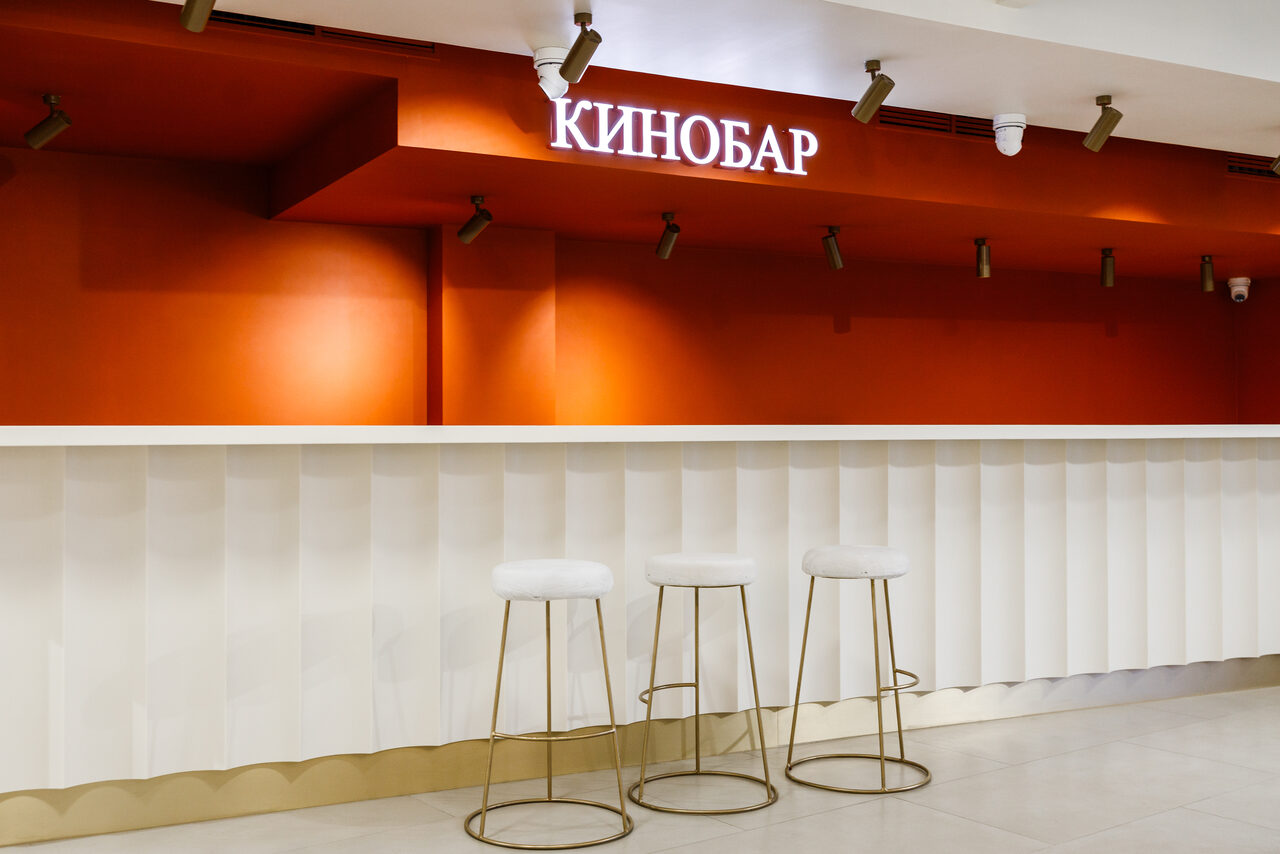 Арендовать Горький Холл. Барная стойка с рифлёной белой панелью и тремя барными стульями с белой обивкой и золотыми ножками. На фоне оранжевая стена с надписью "Кинобар" и установленными светильниками. Потолок белый с встраиваемыми светильниками.