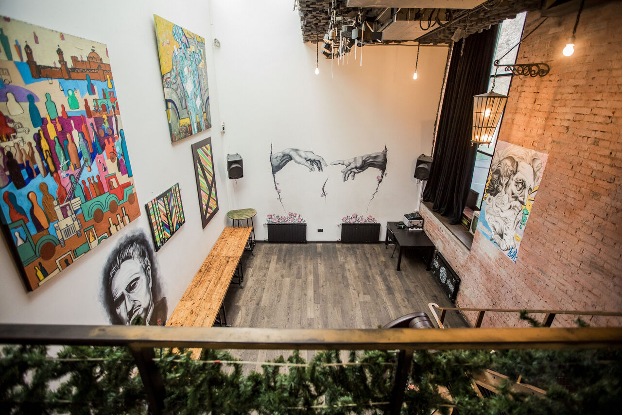 Арендовать Street Art Loft (Стрит Арт Лофт). Лофт-пространство с высокими потолками, стенами из белого кирпича и декором в виде граффити и картин. Деревянный пол, скамейки, столы, подвесное освещение, лестница. На стене розовый кирпич, черные шторы, нетекстовые знаки.