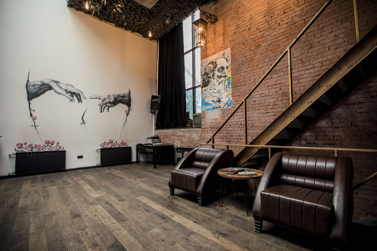 Арендовать Street Art Loft (Стрит Арт Лофт). Лофт-пространство с кирпичными стенами и деревянным полом. Два кожаных кресла и круглый столик. На одной стене граффити, на другой — картина. Металлическая лестница и большие окна с тяжелыми шторами. Высокие потолки, точечное освещение.