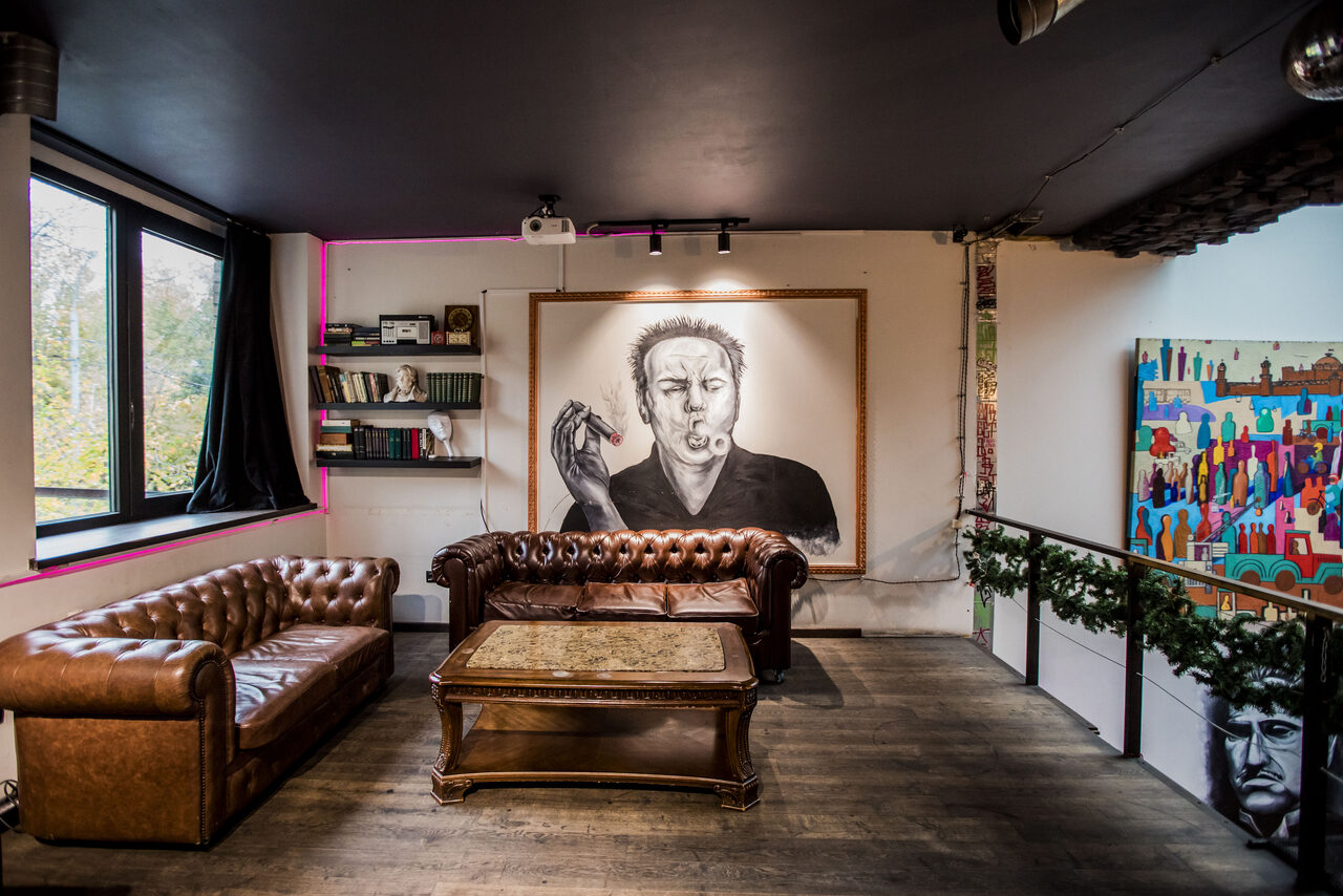 Арендовать Street Art Loft (Стрит Арт Лофт). Лофт-пространство с кожаными диванами и деревянным столиком на фоне портрета на стене, книжные полки, большое окно, яркая картина справа, пол из тёмного дерева, чёрный потолок, проектор, светильники, зелёный декор на перилах.