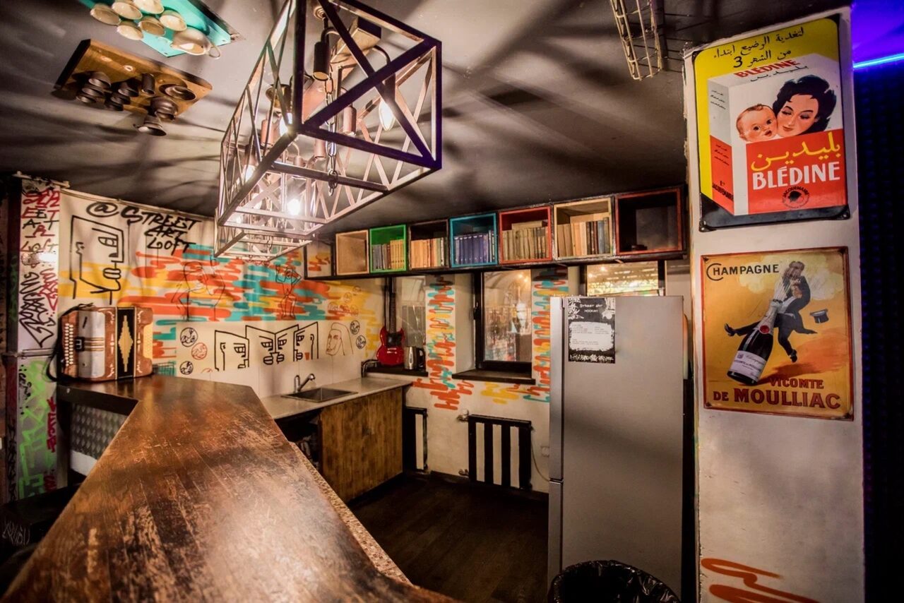 Арендовать Street Art Loft (Стрит Арт Лофт). Барное пространство с деревянной стойкой и стеллажами. Стены украшены граффити и постерами с надписями. Плиточные стены бежевого и разноцветного цвета. Металлические элементы освещения. Окна с черными рамами. Холодильник серого цвета.