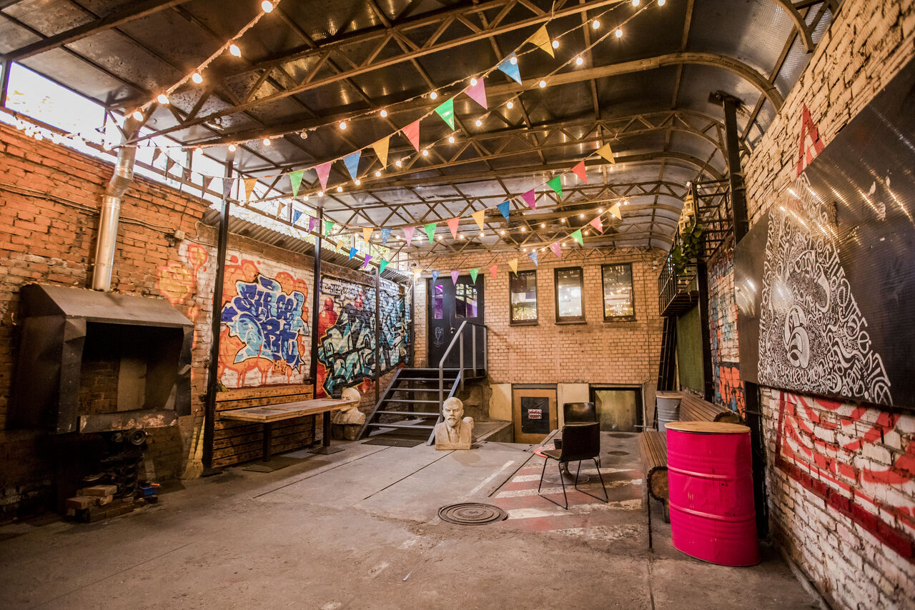 Арендовать Street Art Loft (Стрит Арт Лофт). Промышленное пространство с кирпичными стенами, граффити, металлической крышей. На полу микс брусчатки и плитки, разноцветные флажки и гирлянды. Имеется камин, деревянная скамья, розовая бочка, стул. Видна лестница и двери.