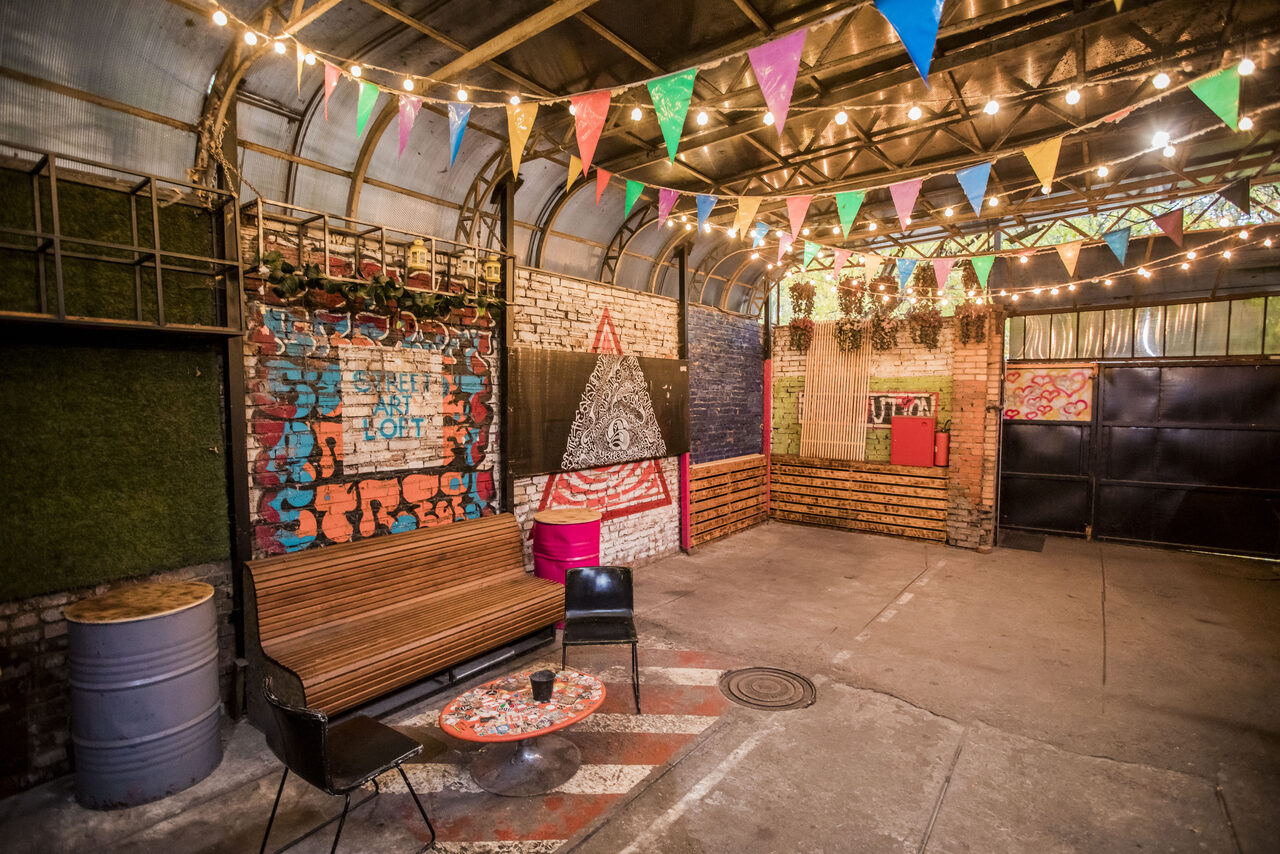 Арендовать Street Art Loft (Стрит Арт Лофт). Промышленное пространство с кирпичными стенами, зоной с деревянной скамейкой и круглыми столиками, подвесными разноцветными флажками и гирляндами ламп. Металлические конструкции и зелёные декоративные элементы на стенах.